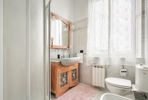 A bathroom with a wooden vanity, a white sink, a mirror, a toilet, and a window with lace curtains.