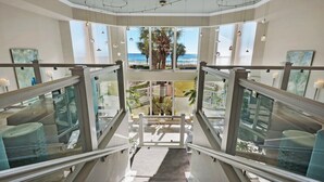 A modern hotel interior with a glass staircase leading to a balcony with a view of the ocean and palm trees. - Silver Beach Towers West 1105 | Destin | Beachfront, Outdoor Pool | My Beach Getaways (Destin)