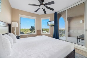 A hotel room with a large bed, ceiling fan, TV, and a balcony with a view of palm trees.
