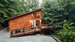 A wooden cabin with a deck and a stone chimney, surrounded by dense forest. - Outside Inn | Hocking Hills, Hot Tub, Views (Sugar Grove)