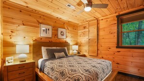 A wooden cabin bedroom with a bed, nightstands, a ceiling fan, and framed artwork on the wall. - Outside Inn | Hocking Hills, Hot Tub, Views (Sugar Grove)