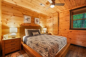 Un dormitorio de cabaña de madera con una cama, mesitas de noche, un ventilador de techo y cuadros en la pared.