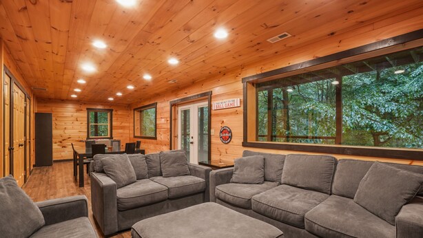 A wooden interior with a sofa set, a dining table, and a large window overlooking a forest. - Outside Inn | Hocking Hills, Hot Tub, Views (Sugar Grove)