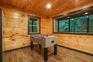 Habitación con piso de madera, mesa de fútbol de mesa y dos ventanas con vista a un bosque.