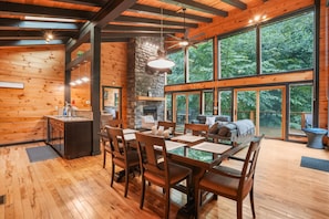 A spacious dining area with a large wooden table and chairs, a stone fireplace, and large windows offering a view of the surrounding forest.
