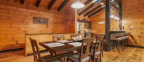 A wooden interior with a dining area featuring a table and chairs, a kitchen area with appliances, and a bar with stools.