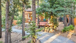 A rustic cabin surrounded by tall trees, with a paved driveway leading to a wooden deck. - The Nest on Towne Trail Big Bear Lake - Hot Tub - Village - (Big Bear Lake)