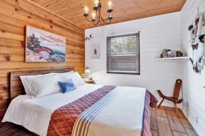 Ein Schlafzimmer mit Holzbalkendecke, einem Bett mit weißer Bettwäsche und einer Steudecke, einer Kronleuchter, einem Fenster mit Jalousien sowie einem Regal mit Büchern und Dekoartikeln.