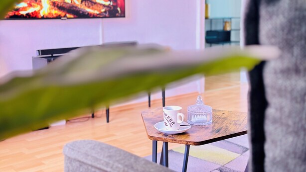 Ein gemütliches Wohnzimmer mit einem Holztischchen, einer weißen Tasse und einem Glasbehälter. Ein Fernseher zeigt ein Lagerfeuer und ist an der Wand befestigt.