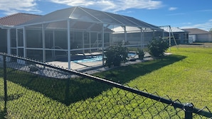 A covered patio area with a pool, surrounded by a chain-link fence and greenery.