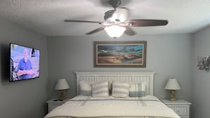 A bedroom with a bed, two bedside tables, a ceiling fan, a TV, and a painting on the wall.