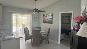 A spacious kitchen with a dining area, featuring a ceiling fan, a dining table with chairs, and a view of the outdoors through large windows.