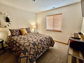 A bedroom with a bed, a bedside table, a lamp, a mirror, and a window with blinds.