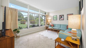 Ein großzügiges Wohnzimmer mit einem großen Fenster, einem türkisfarbenen Sofa, einem Holztisch und einem Fernseher auf einem Holzgestell.