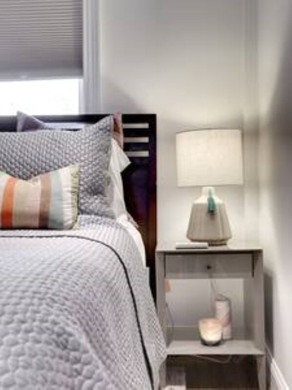 A neatly made bed with a grey quilt, a bedside lamp, and a small bedside table with a lamp and a cup.