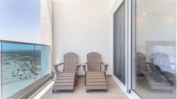 A balcony with two Adirondack chairs, a view of a beach, and a glimpse of a bedroom with a bed and a nightstand.
