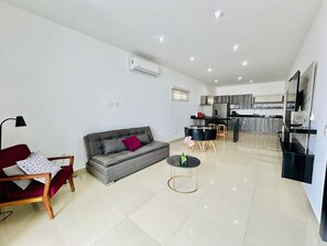 Uma sala de estar moderna com um sofá cinza, uma poltrona vermelha e uma mesinha de centro. Ao fundo, há uma cozinha com mesa e cadeiras.