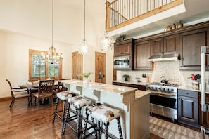 A kitchen with wooden cabinets, a central island, bar stools, a chandelier, and a view of the outdoors.