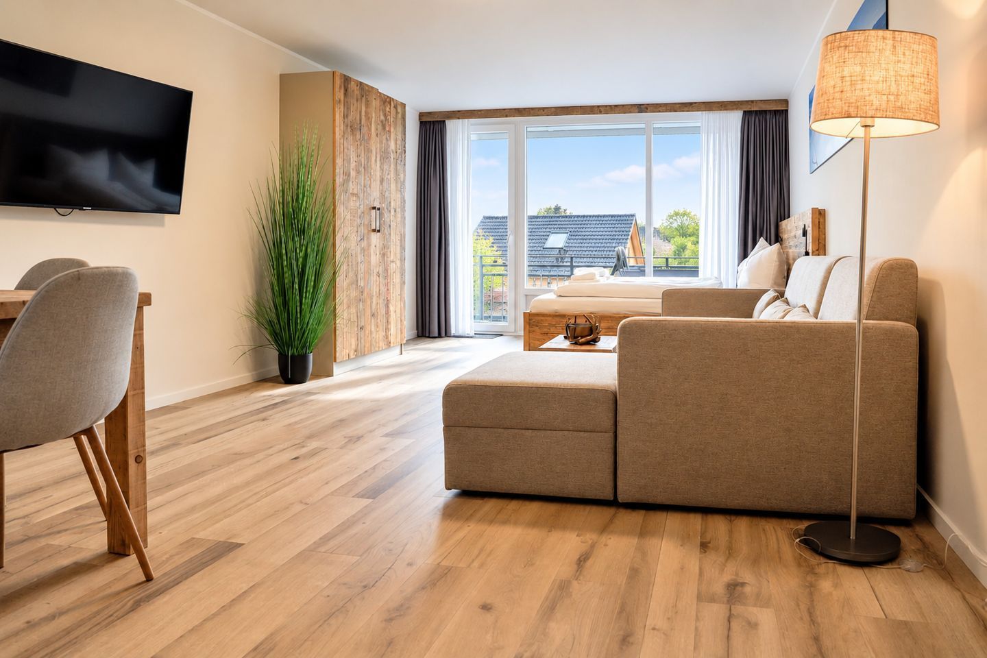 A modern living room with a sofa, a dining table with chairs, a TV mounted on the wall, and a large window with a view of buildings.