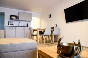 Un espace de vie moderne et compact comprenant un coin cuisine, un coin repas avec quatre chaises, un téléviseur à écran plat fixé au mur et un lit.