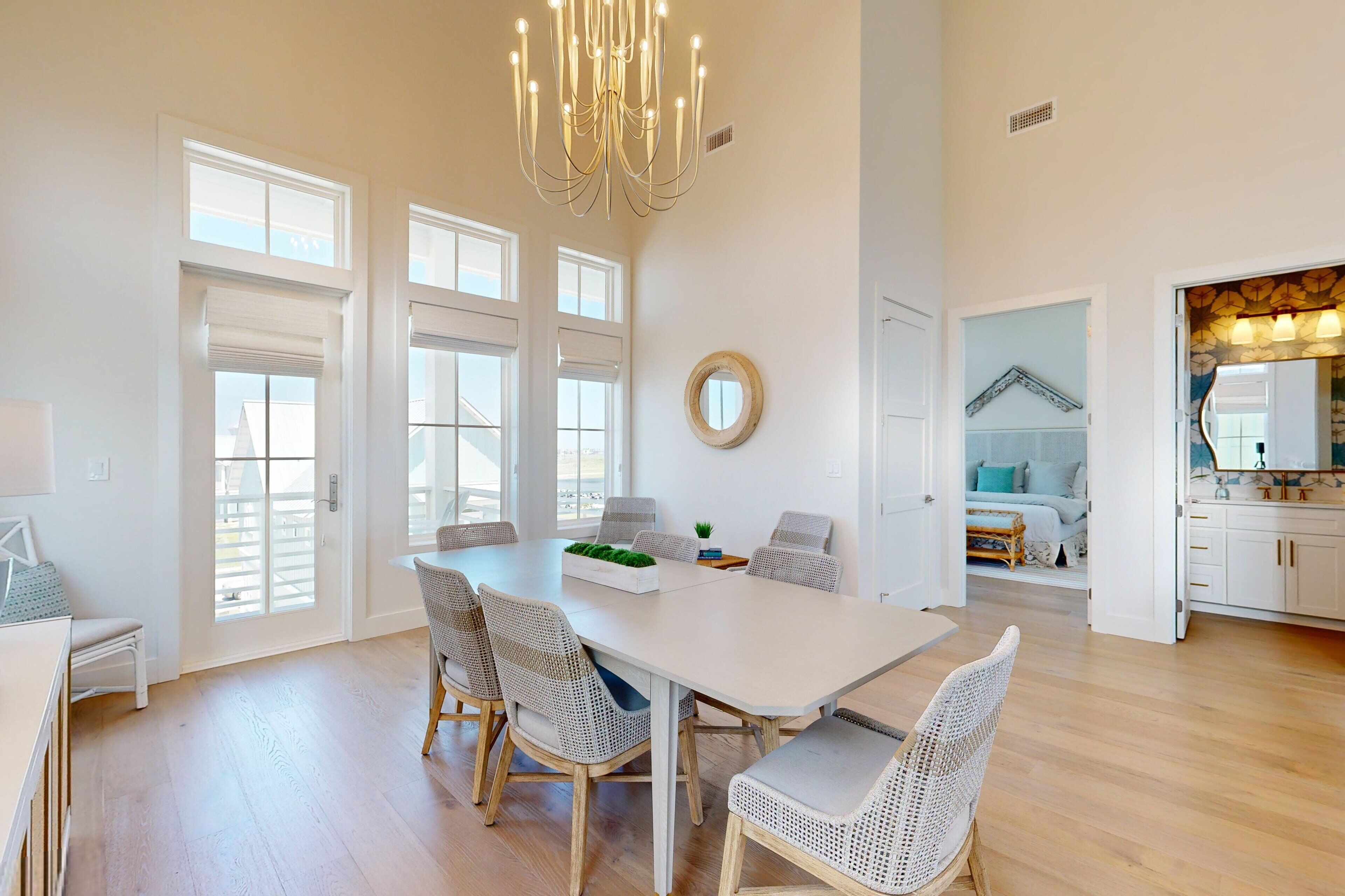 A spacious dining area with a large table and wicker chairs, a chandelier, and large windows allowing natural light.