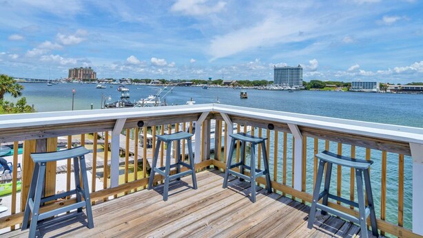 Una terraza de madera con tres taburetes azules que dan a un puerto deportivo con barcos y edificios al fondo.