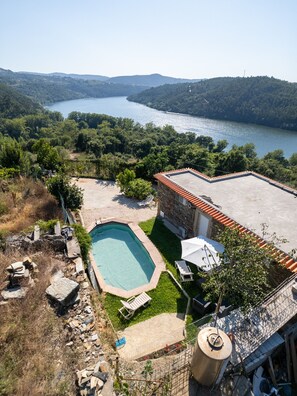Un espace piscine agrémenté d’une maison en pierre, avec vue sur un lac et entouré de verdure.