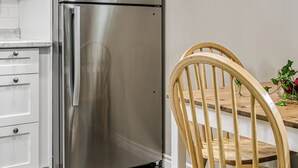 Een keuken met witte kastjes, een roestvrijstalen koelkast, een houten eettafel met stoelen en een plant in een pot bij het raam.