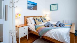 A bedroom with a wooden bed, white bedding, two pillows with 'Ocean Days' text, a beach photo on the wall, and a small bedside table with a lamp. - Sea Glass - 2 Bedroom Apartment - Manorbier (Tenby)