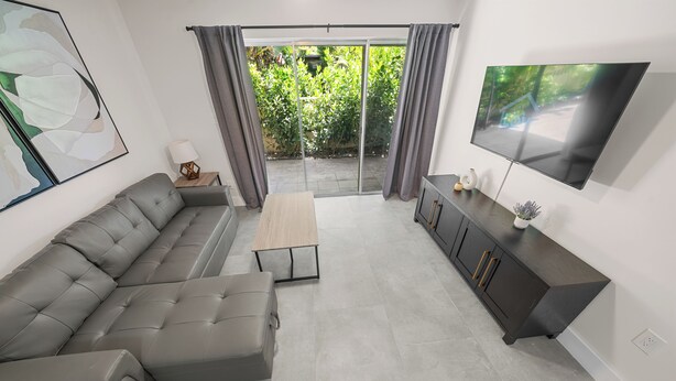 Una sala moderna con un sofá seccional gris, una mesa de centro, un televisor en la pared y un ventanal corredero que da a un jardín.