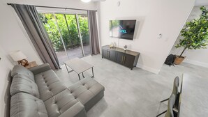 Una sala de estar moderna con un sofá seccional gris, un televisor de pantalla plana, un ventanal corredero que da a un jardín y una decoración de estilo minimalista.