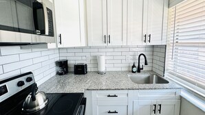 Cocina compacta con armarios blancos, pileta de acero inoxidable, microondas, tetera y cafetera.