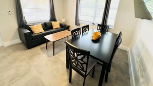 Una sala de estar moderna con un sofá, una mesa de comedor y sillas. Hay un ventilador de techo y una ventana con cortinas.