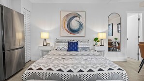 Une chambre moderne avec un lit, des lampes de chevet, un grand tableau abstrait, un miroir et un coin repas visible à travers une porte ouverte.