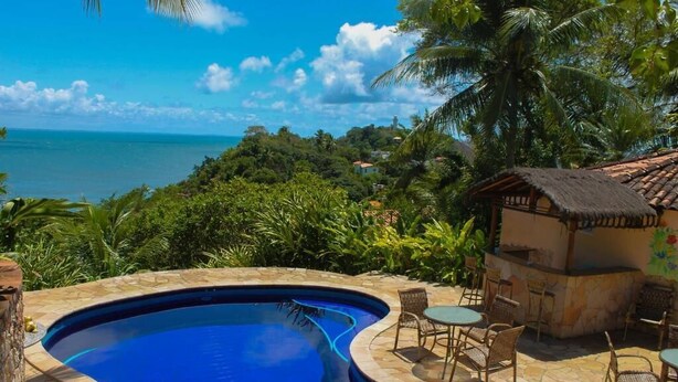 A swimming pool with a curved edge, surrounded by stone tiles and wicker furniture. - Pousada Colibri (Cairu)