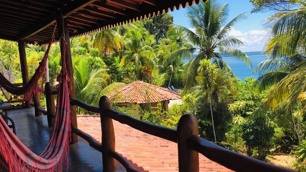 A hammock under a wooden pergola overlooking a lush garden with palm trees and a view of the sea. - Pousada Colibri (Cairu)