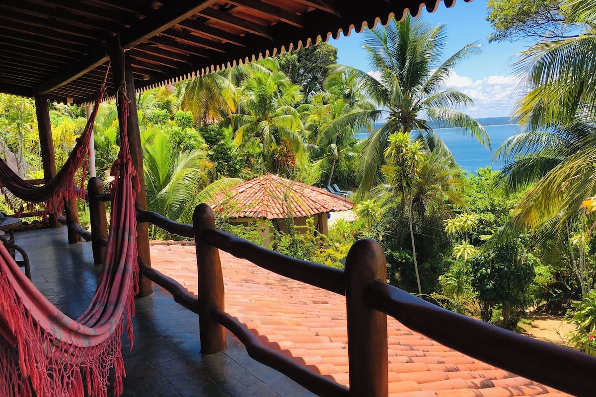 Un hamac suspendu sous une pergola en bois, donnant sur un jardin luxuriant, avec des palmiers, et offrant une vue sur la mer.