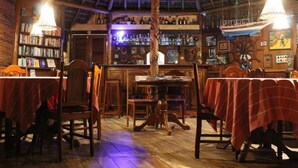 A restaurant with wooden tables and chairs, a bar area with bottles, and a thatched roof. - Pousada Colibri (Cairu)