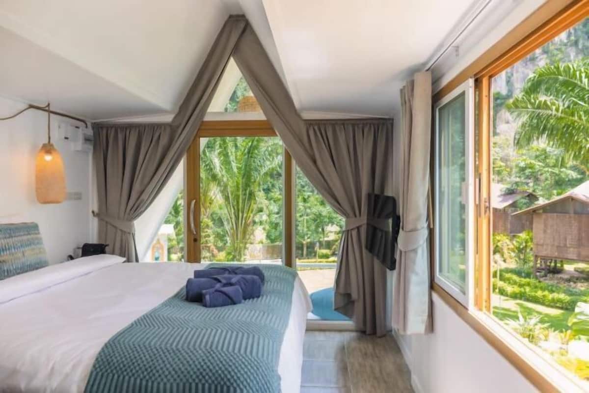 Ein Schlafzimmer mit Bett, Vorhängen, Blick ins Grüne und einem Fenster.