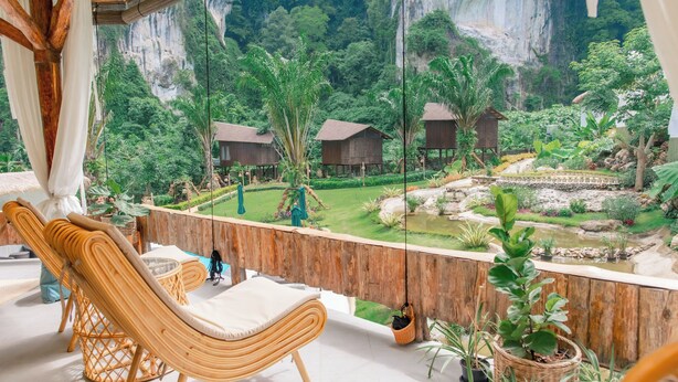 A balcony with wicker furniture, a view of a lush garden, and wooden structures in the background.