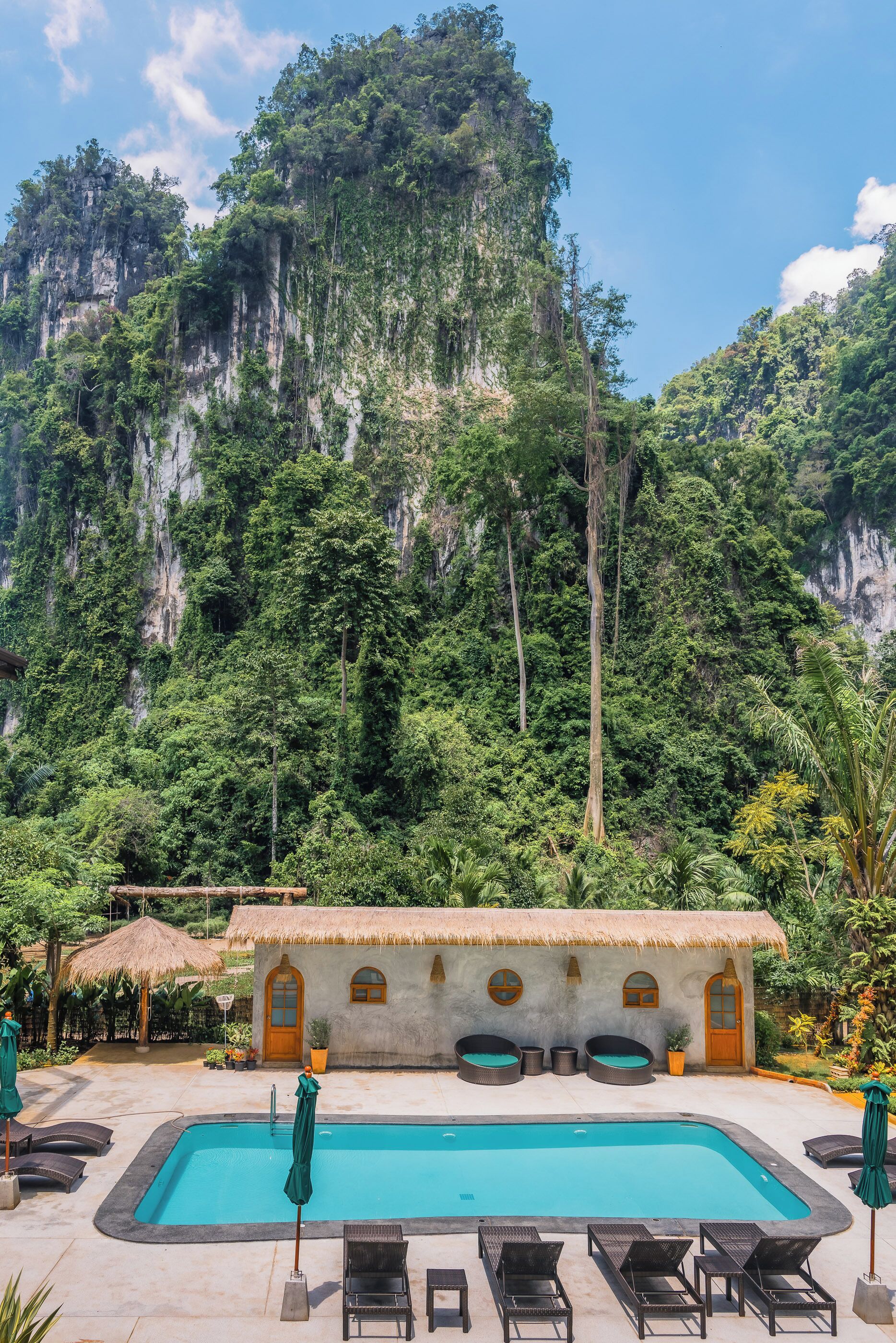 A pool area with lounge chairs and a thatched-roof building, surrounded by lush greenery and a cliff.