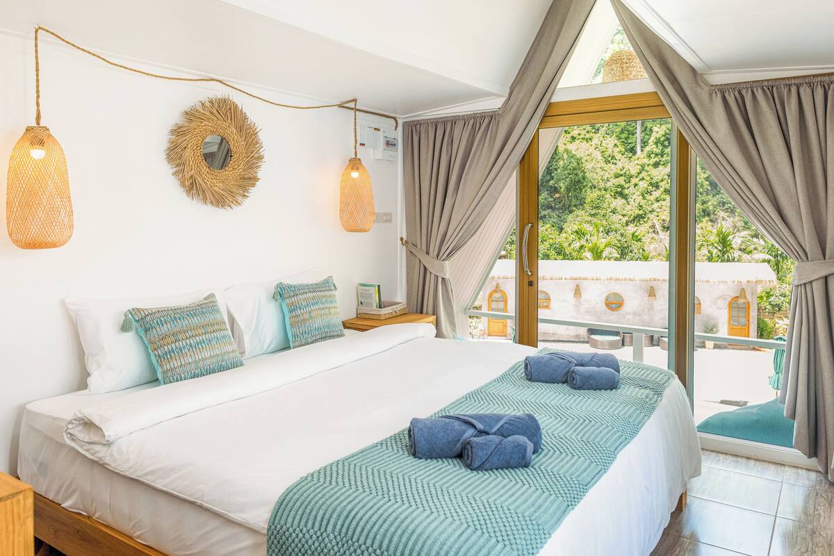 Ein Schlafzimmer mit einem großen Bett, zwei Wandlampen, einer gewebten Wanddekoration und Blick auf einen Innenhof durch eine Glastür.