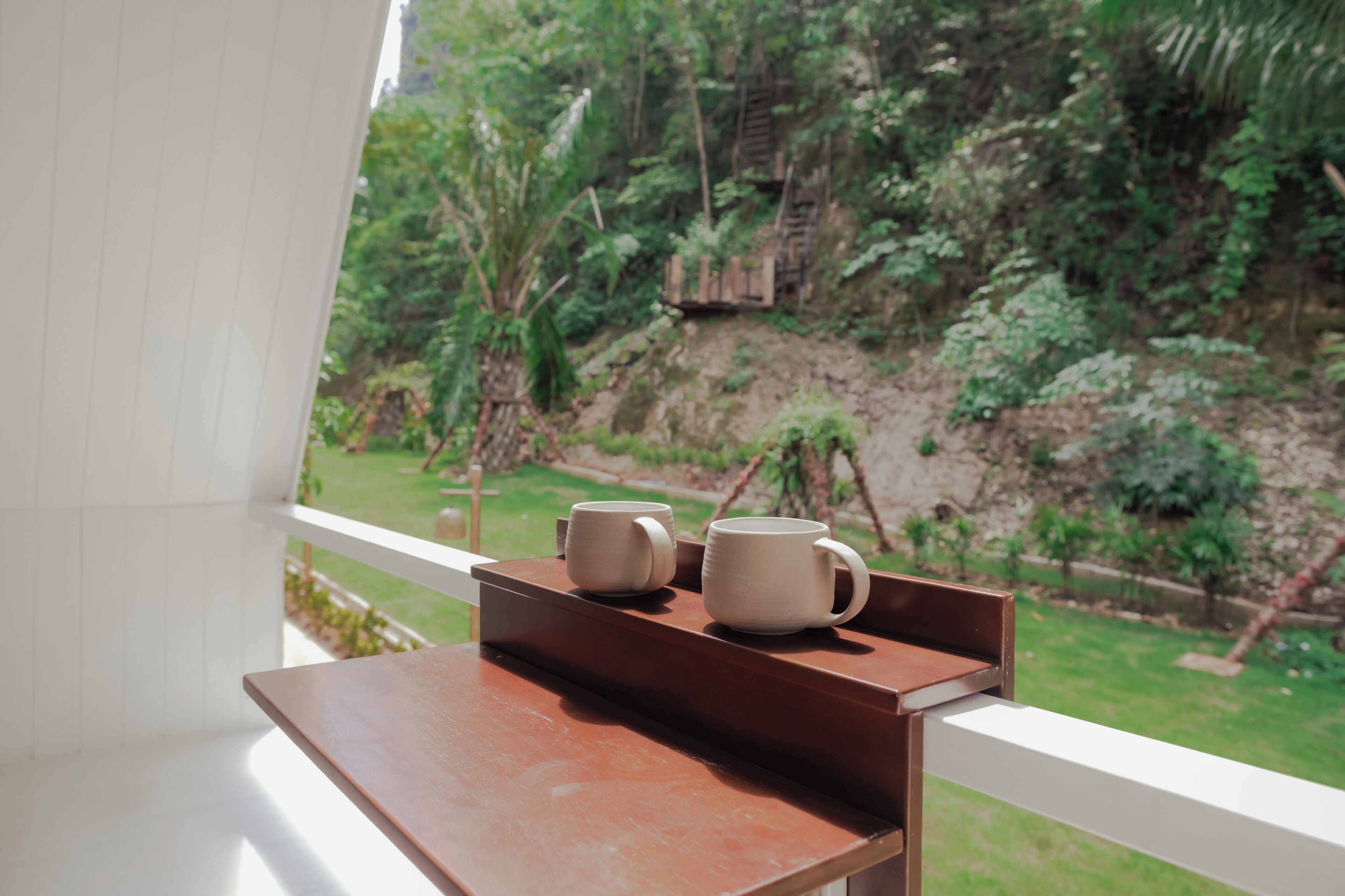 Un balcon avec une table en bois sur laquelle sont posées deux tasses blanches, donnant sur un jardin luxuriant avec un escalier en bois.