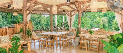 An open-plan dining area with wooden tables and chairs, surrounded by wicker furniture and potted plants.