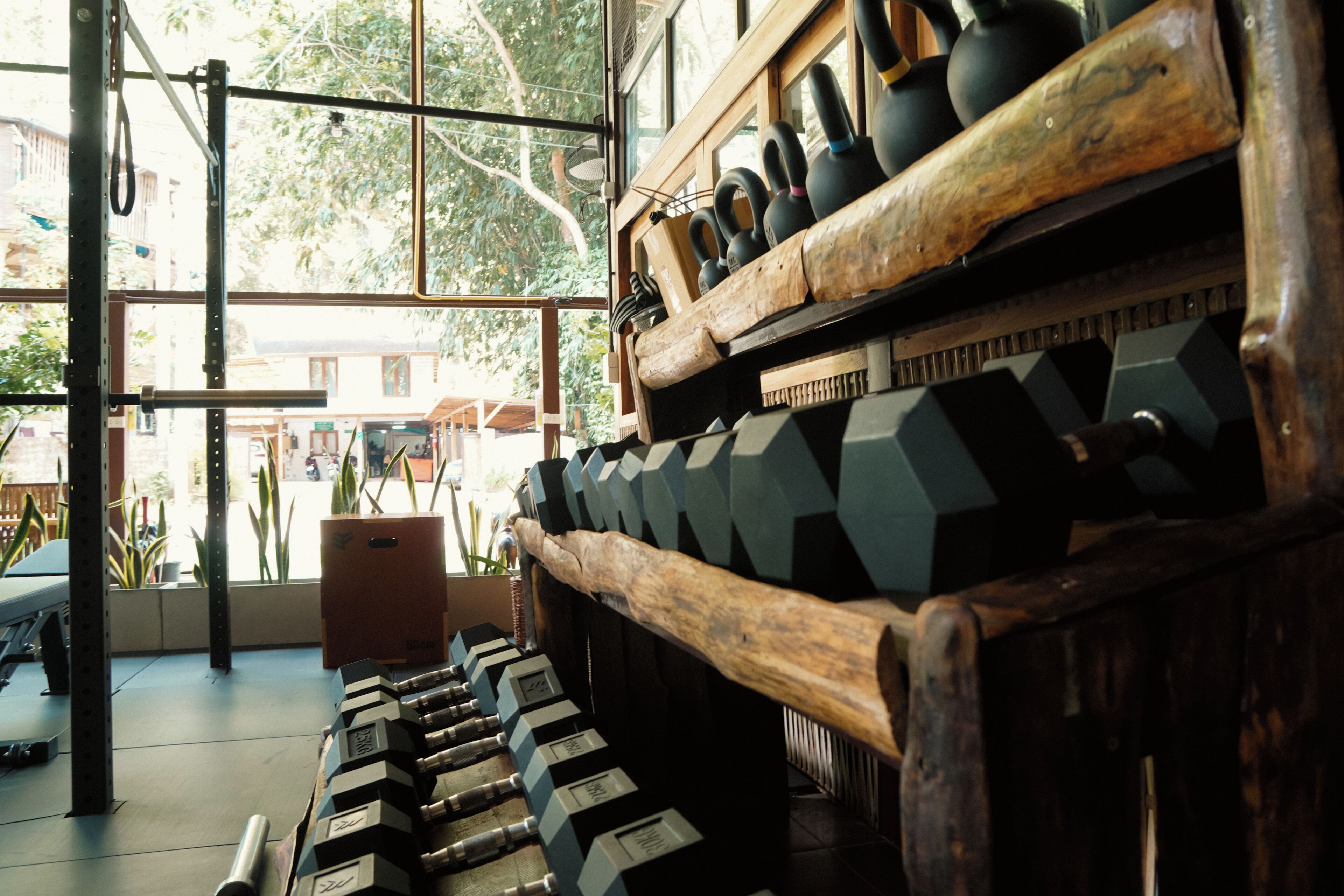 A gym with kettlebells and dumbbells arranged on racks, large windows, and a view of the outside.