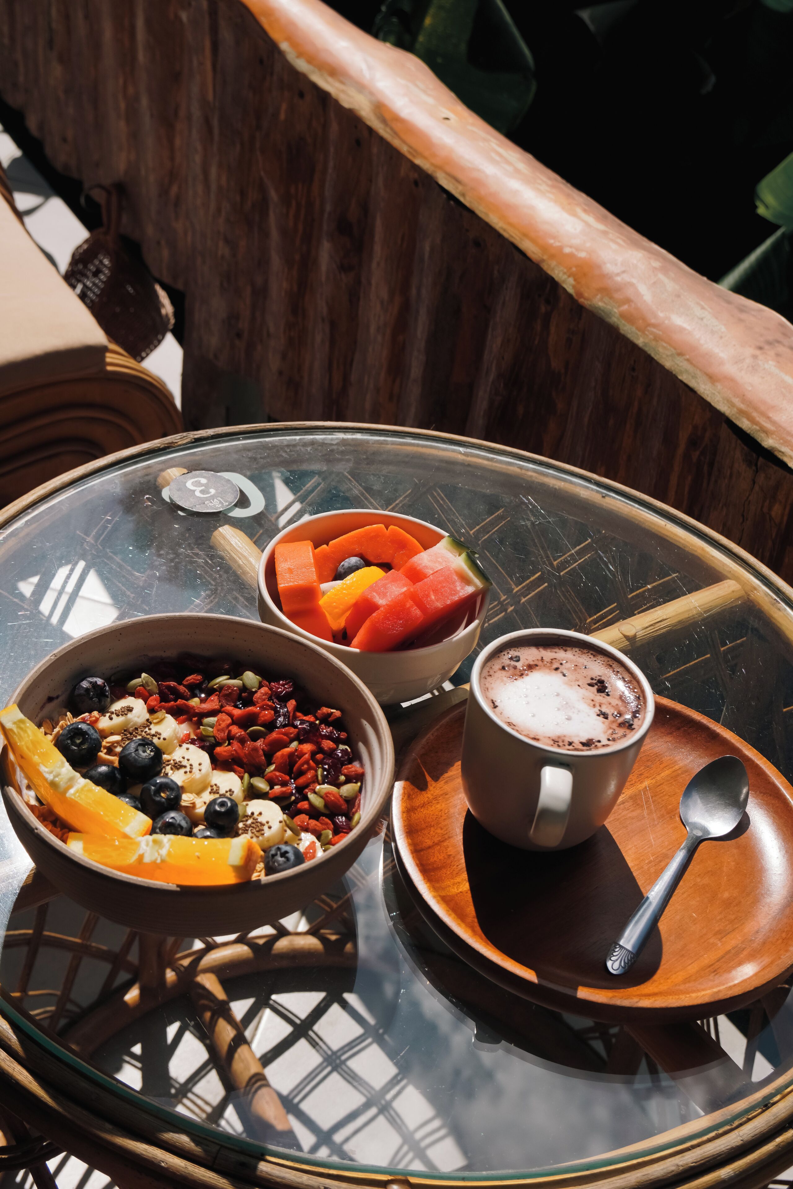 A glass table with a bowl of fruit, a cup of coffee, and a spoon.