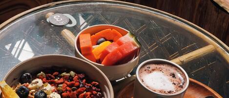 A glass table with a bowl of fruit, a cup of coffee, and a spoon.