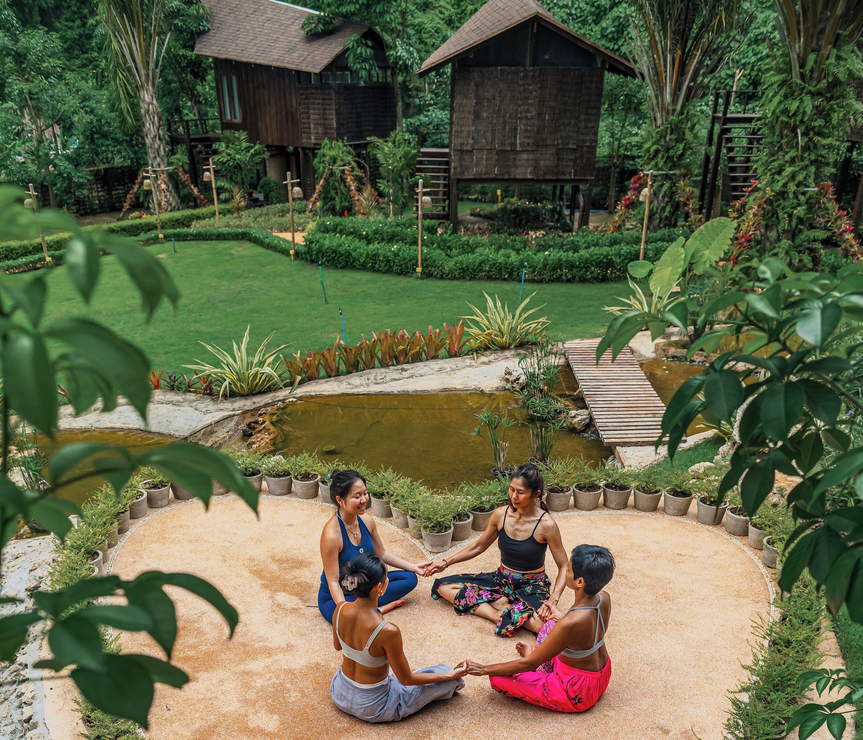 A group of people sitting in a circle on a sandy area surrounded by potted plants and a small pond, with wooden structures and lush greenery in the background.