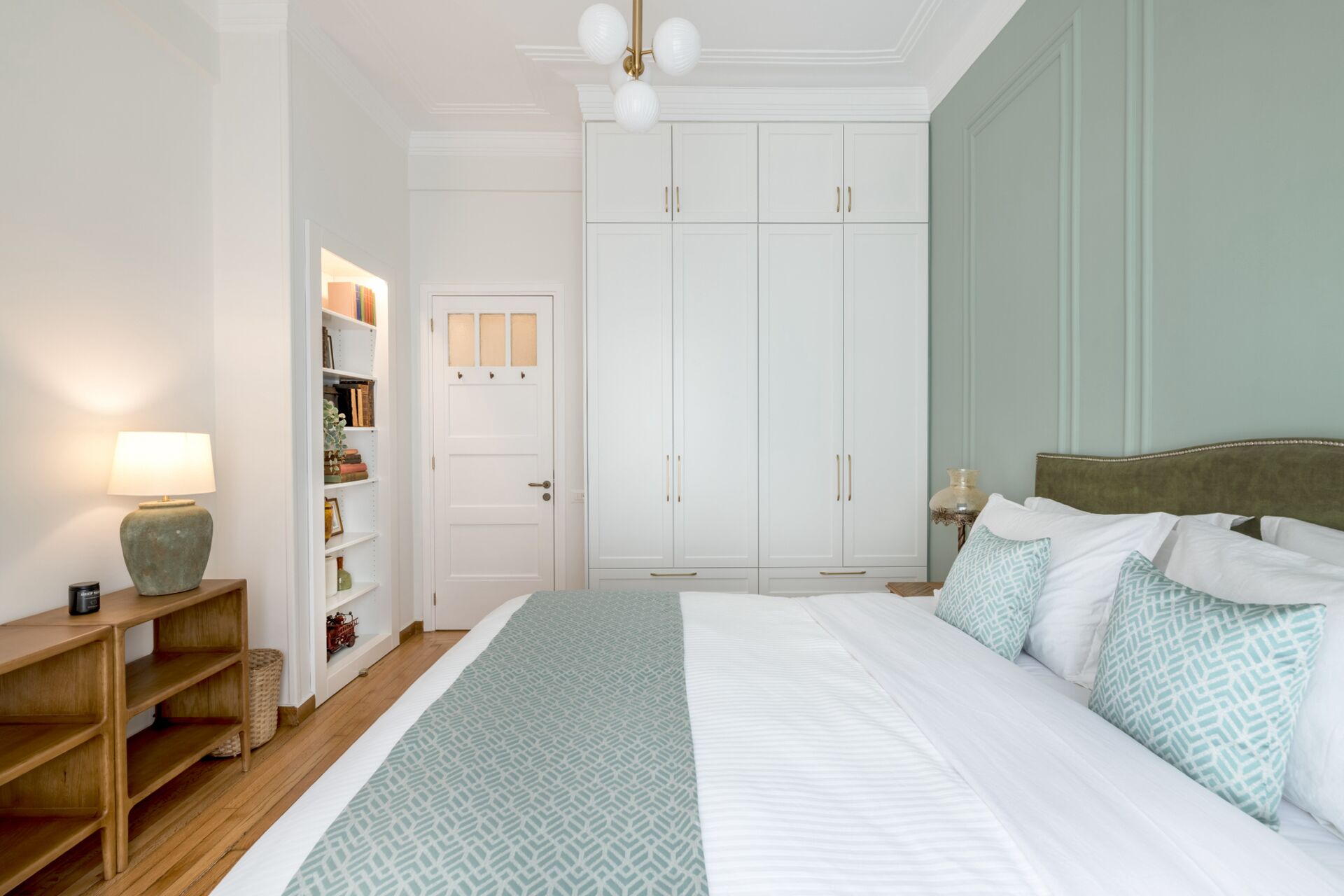 A bedroom with a bed, a nightstand, a lamp, a wardrobe, and a shelf with books and decorative items.
