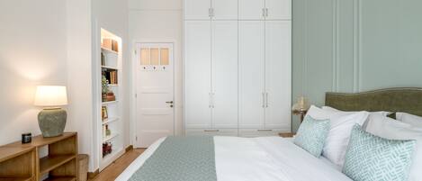 A bedroom with a bed, a nightstand, a lamp, a wardrobe, and a shelf with books and decorative items.